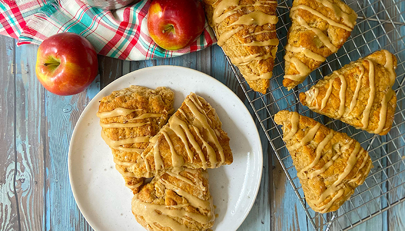 An aerial view shows a plate of scones next to more scones on a wire rack. A tea towel, red apples, and a cup of tea are visible.