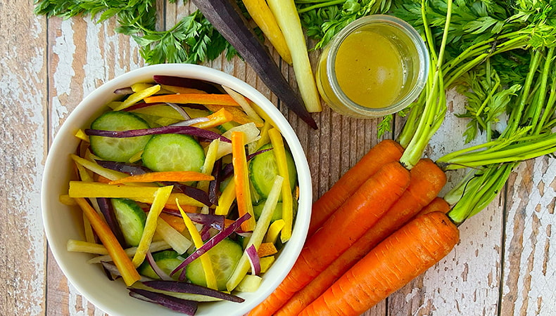 Une vue aérienne montre un petit bol de salade de carottes et de concombres sur une table en bois usée par le temps. Des carottes orange et arc-en-ciel ainsi qu'un petit pot de sauce à salade sont également visibles.