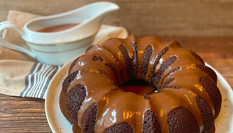 A chocolate bundt cake sits on a platter. A serving dish with chocolate sauce is visible in the background.