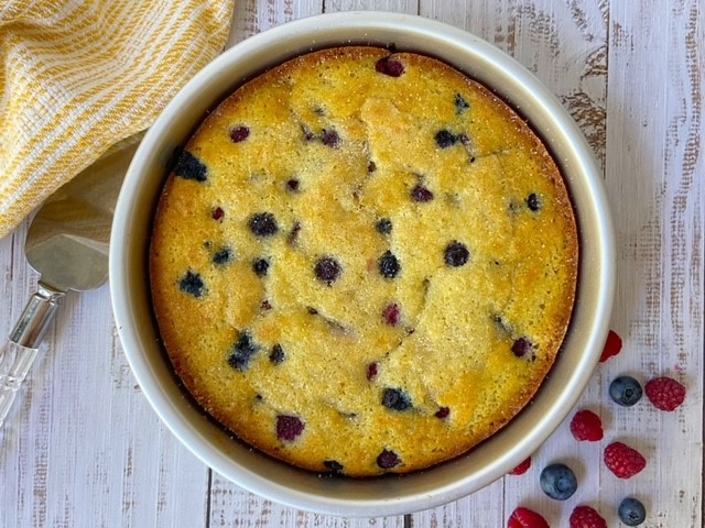 An aerial view shows a Berry Buttermilk cake in a round pan, sitting on top of a faded wooden surface. Assorted berries are sprinkled next to the pan.