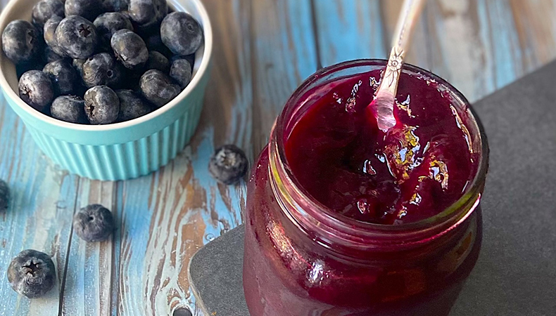 Une sauce violet foncé repose dans un pot ouvert avec une cuillère qui en ressort. Un petit bol de bleuets se trouve près du pot, sur une surface de bois.