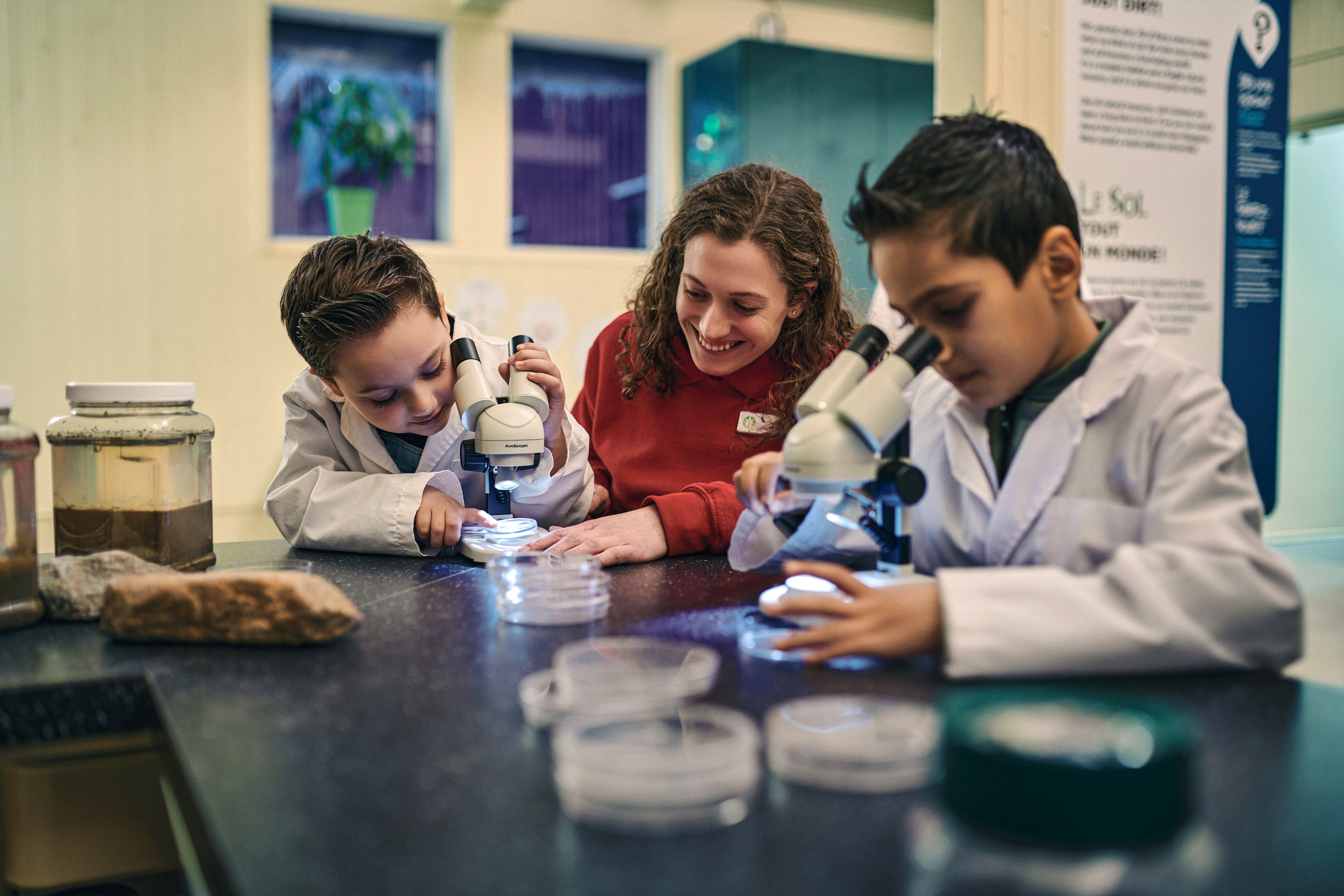 Deux enfants en blouse blanche regardent des boîtes de Petrie dans des microscopes. Un employé du musée aux longs cheveux bruns et au polo rouge leur sourit. Au premier plan, on aperçoit un pot de terre et d'autres boîtes de Petrie.