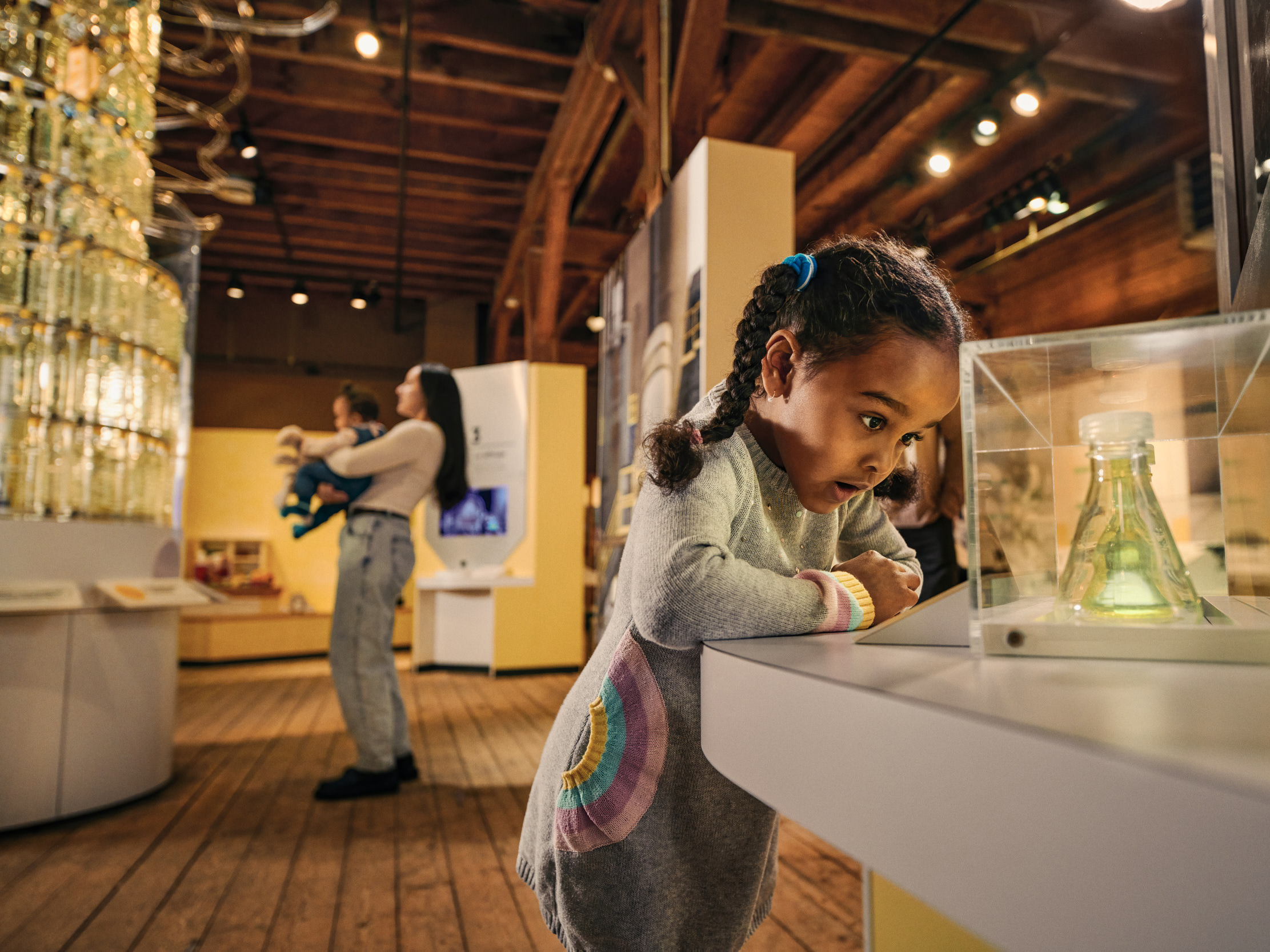 A young child with a braid looks in awe at a beaker of light yellow liquid, enclosed in a plexiglass case. In the background, a woman and baby look on at a museum exhibition.