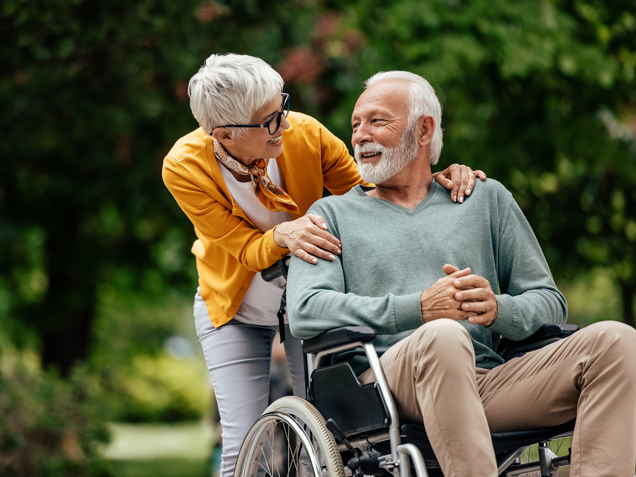 Un homme âgé à la barbe blanche est assis dans un fauteuil roulant. Une femme aux cheveux blancs se tient derrière lui, lui tenant affectueusement les épaules.