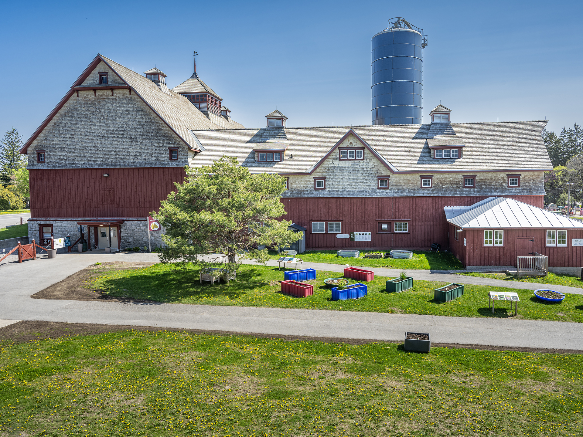 A historic red barn with wooden details, many windows and a blue silo. In the foreground there is a grassy area with planters, a tree and several paved pathways.