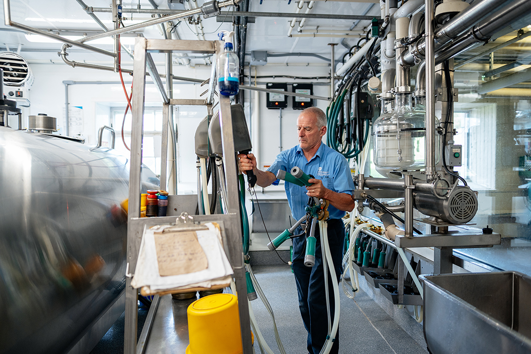 An adult in a blue shirt holds a tube with several attachments. The room is predominately stainless steel, with a large vat to the left and tubing along the right side of the room.