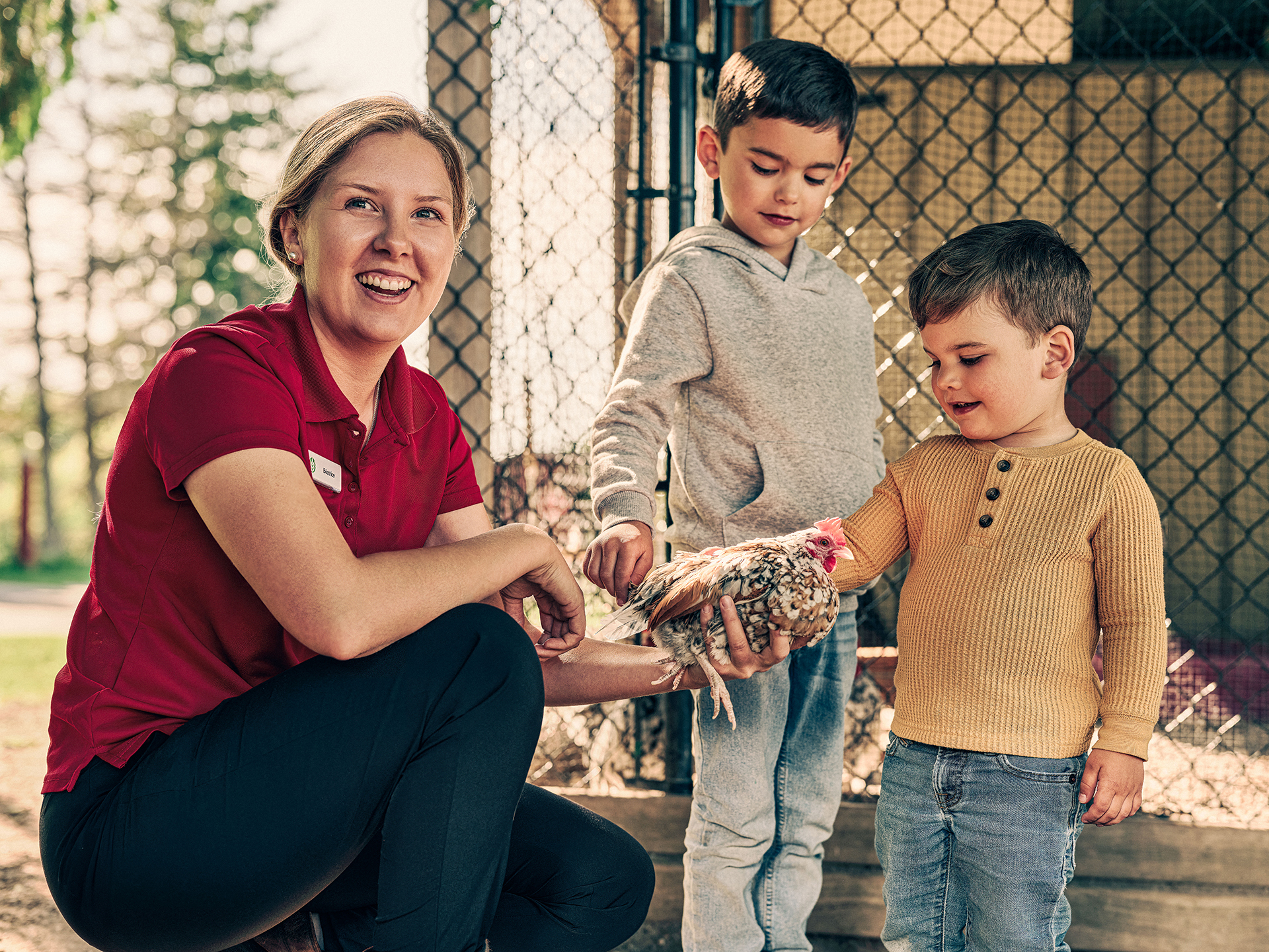 A museum worker with blonde hair and a red shirt holds a small chicken in her hand. Two young children in sweaters and jeans look on intently, reaching out to touch the chicken’s brown, black and white mottled feathers.