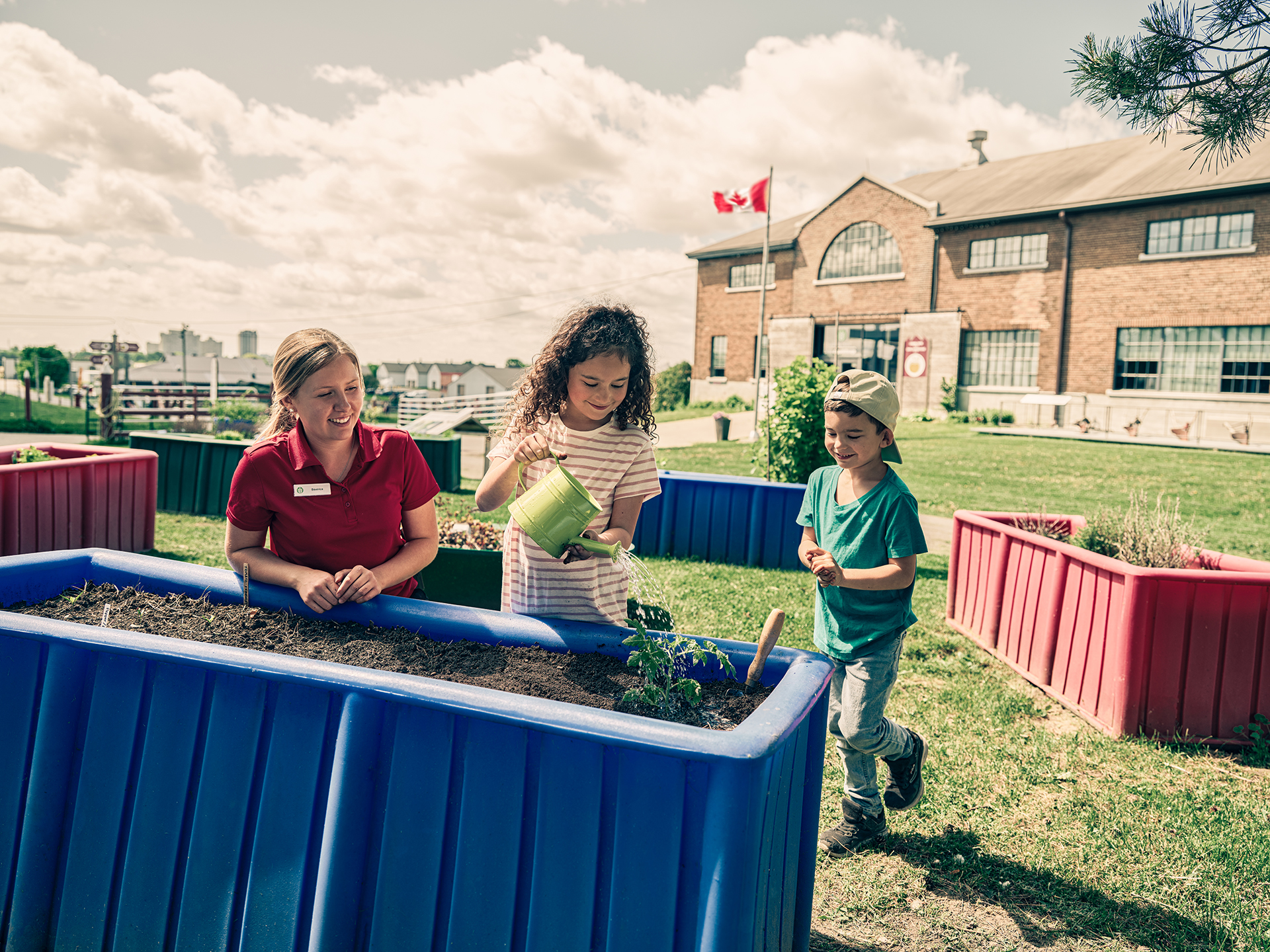 There are several colourful waist-high garden beds outdoors. A child uses a green watering can to water a plant. An adult and another child look on, smiling. The sky is bright and cloudy.