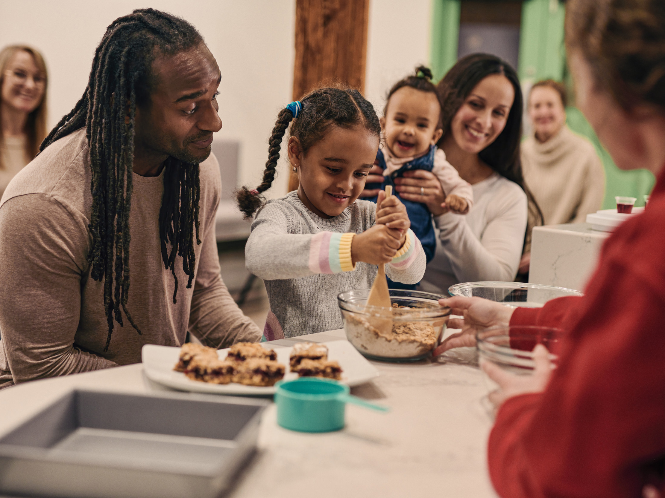 A family participates in a cooking demonstration. A young child stirs ingredients in a bowl with a wooden spoon, while others look on. A plate with desserts and other baking instruments are in the foreground.