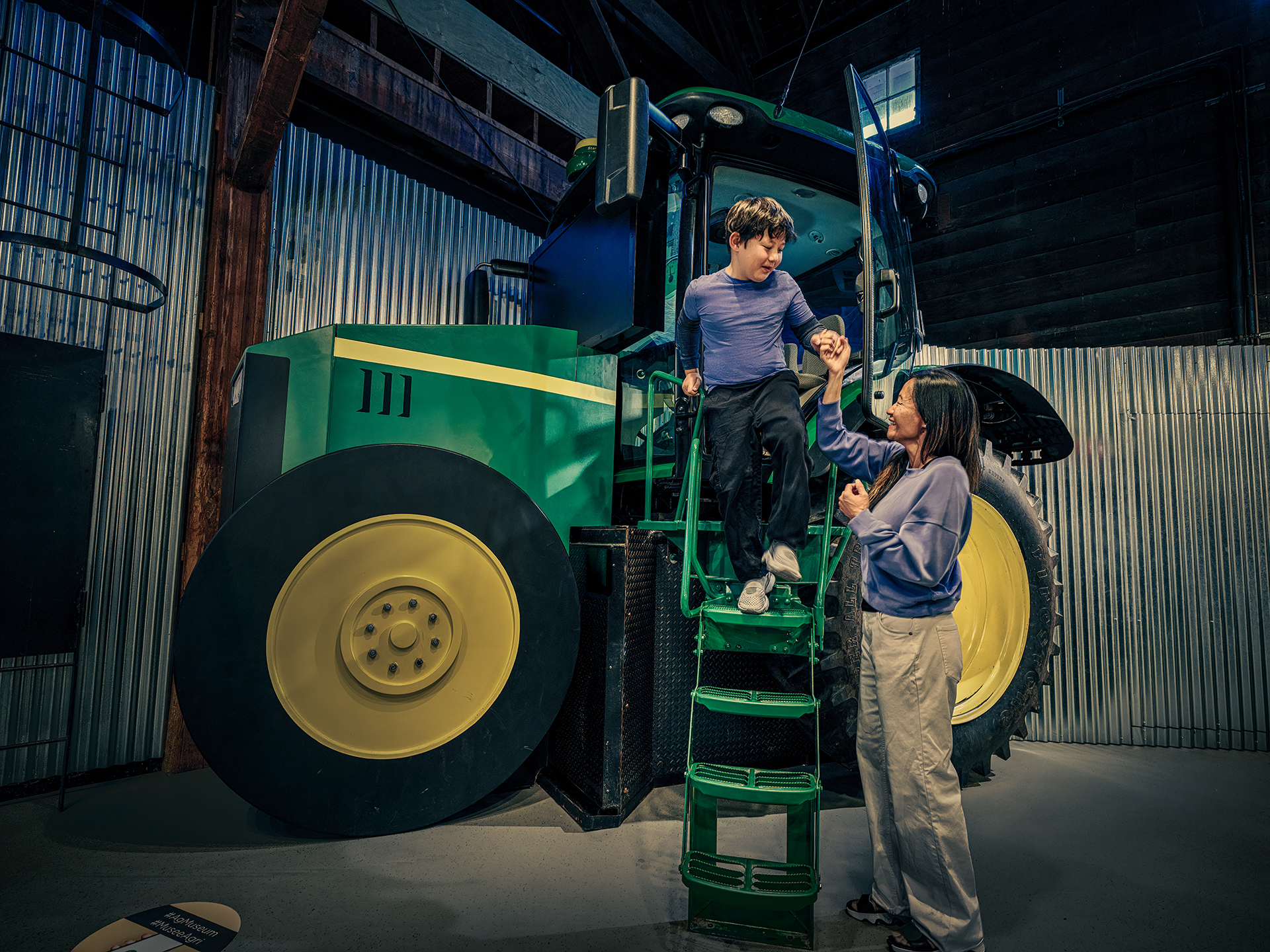 A child holds an adult’s hand as he descends green stairs coming from the cab of a green tractor with large tires and yellow rims. The tractor simulator is indoors at the museum.