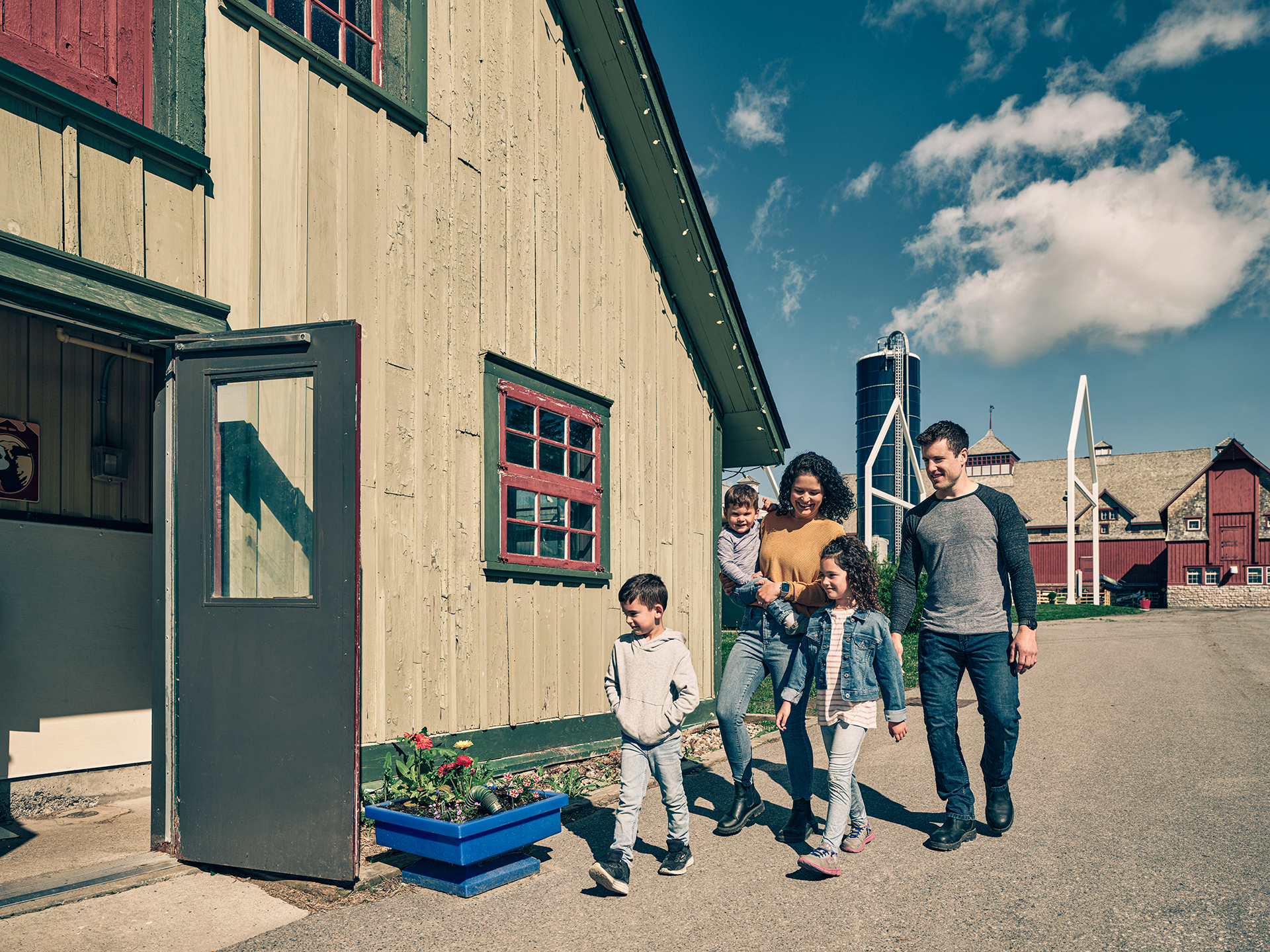 A family of five people – two adults and three children -- walk towards the open door of a light green barn. Another barn, a silo and fluffy clouds are in the background.