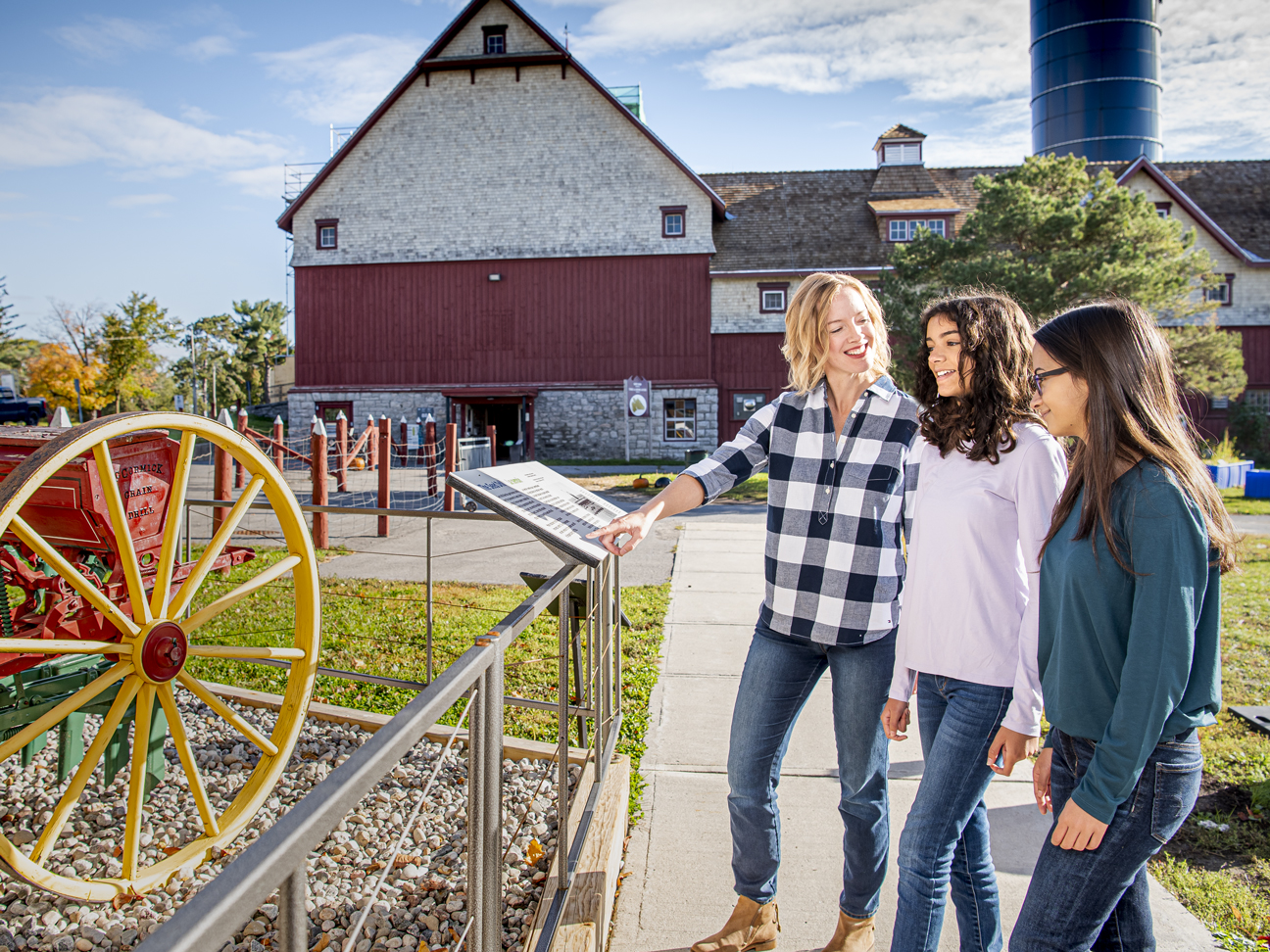 Trois adultes sont réunis en plein air près d'une exposition de musée, regardant du matériel agricole historique avec une grande roue jaune à rayons. À l'arrière-plan, on aperçoit une grande grange rouge. 