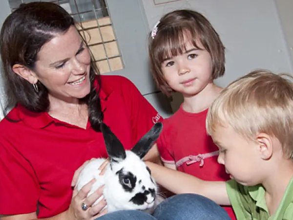 Un guide de musée tient un lapin tacheté blanc et noir. Deux jeunes enfants le regardent.