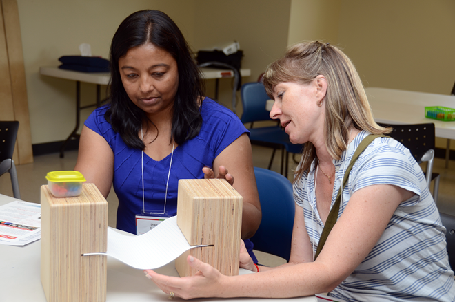 Two teachers sit at a table looking at two large wooden blocks. Set into two slots in the blocks is a white piece of paper.