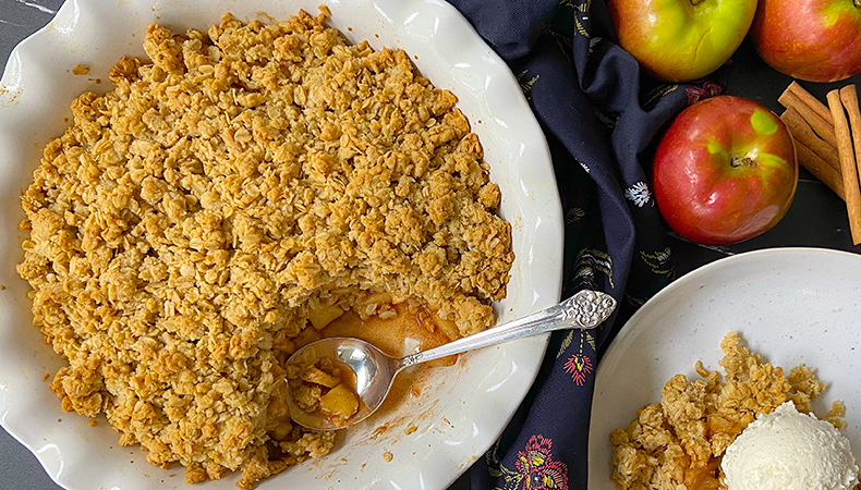 An aerial view shows a large dish of apple crisp, along with a serving size topped with ice cream. Apples and cinnamon sticks are visible next to the dishes.