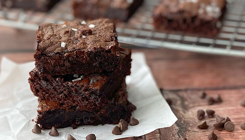 A stack of brownies sit on a wooden surface. In the background, more brownies are arranged on a wire rack.