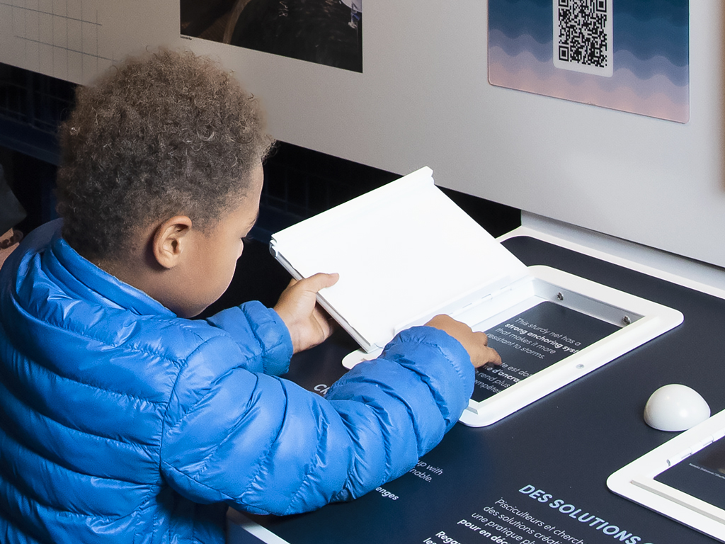 A rear view of a toddler in a blue coat engaging with a Braille panel interactive exhibit at the Canada Agriculture and Food Museum, fostering curiosity and sensory exploration.