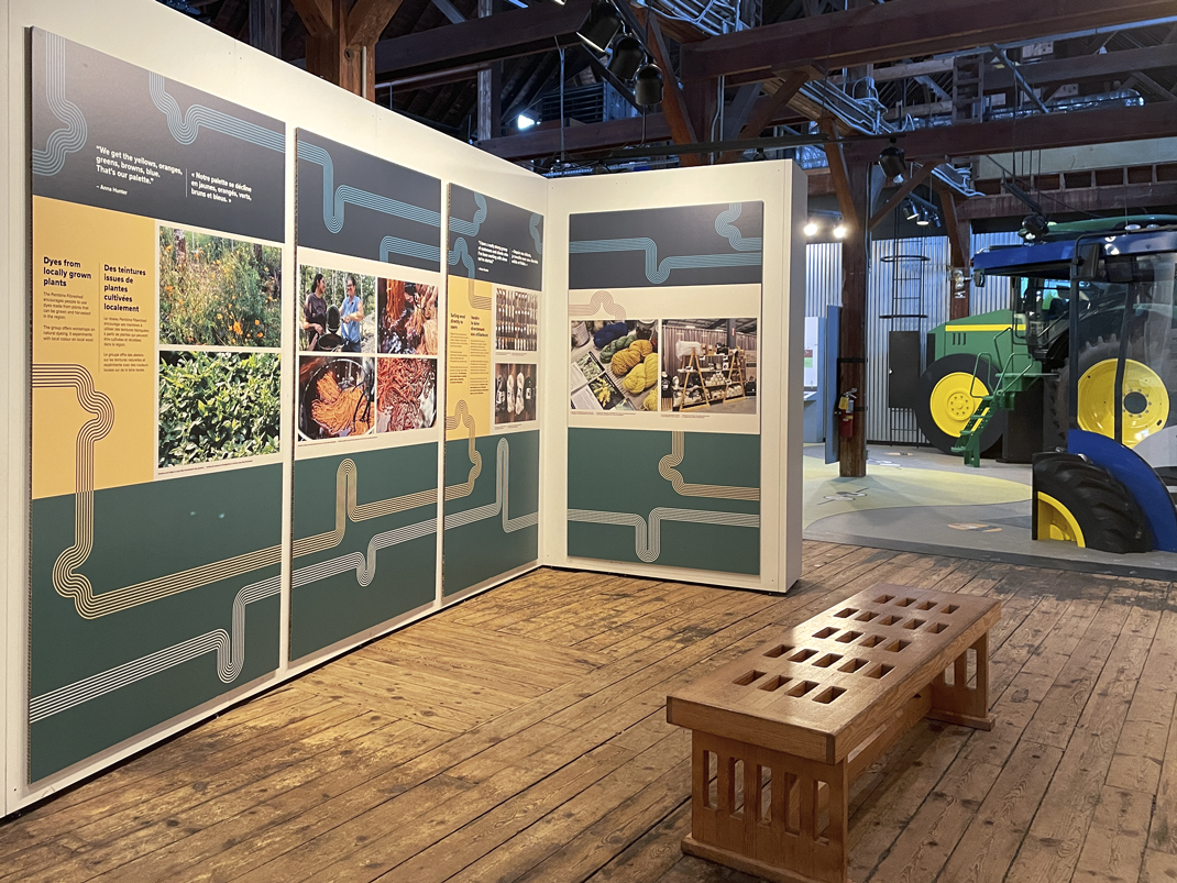 A display in the Sustainable Agriculture Gallery featuring panels with images and text about locally grown plant dyes. The exhibit is surrounded by a wooden floor, a small wooden bench, and a large green tractor in the background.