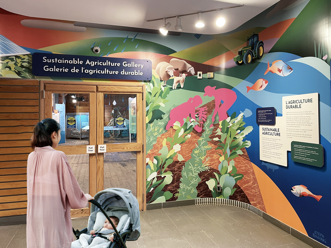 A woman pushes a stroller with a baby as they enter the doors of the Sustainable Agriculture Gallery exhibition. In front of them is a vibrant mural featuring sheep, a tractor, fish, and silhouettes of people cultivating a garden. The display also includes text offering detailed information about the exhibition.