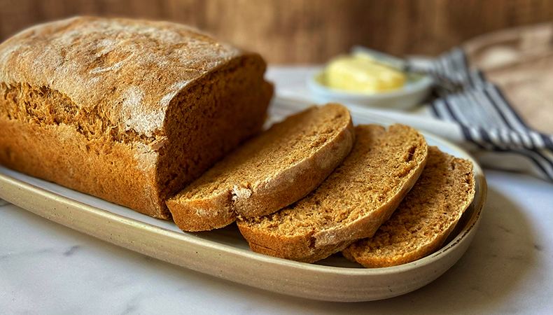 A loaf of bread, with several pieces sliced, sits on a tray on top of a marble counter. A tea towel and a small dish of butter is visible in the background.