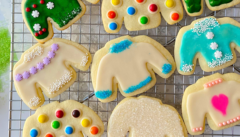 Plusieurs biscuits en forme de chandail d'hiver décorés de couleurs vives sont posés sur un support de séchage de biscuits en fil métallique. Les biscuits sont décorés de bonbons et de glaçage.