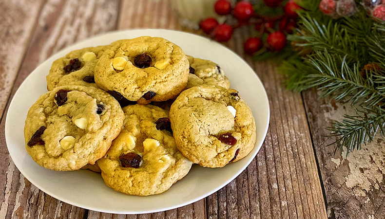 A plate of cookies sits on top of a rustic, wooden surface. A glass of milk and an evergreen branch with red berries are visible next to the plate.