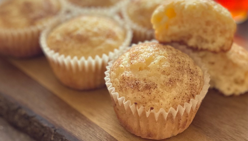 A close-up view of several brown muffins sitting on a wooden surface; a glass jug of maple syrup is visible in the background.