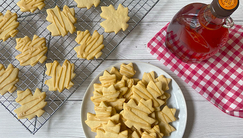 Une vue aérienne montre une étagère en métal et une assiette blanche couverte de biscuits à la feuille d'érable. Une bouteille de sirop d'érable et une serviette à carreaux rouges et blancs sont visibles.