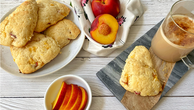 Une vue aérienne montre une assiette de scones sur une surface en bois. Des pêches entières et en tranches ainsi qu'une tasse de café sont posées à côté.