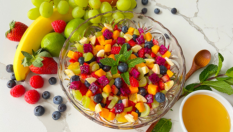 Une vue aérienne montre un bol de salade de fruits coloré sur un comptoir en marbre. Un assortiment de fruits, une cuillère et un petit bol de vinaigrette se trouvent à côté de la salade.