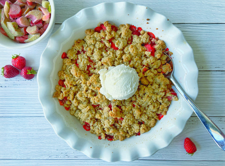 An aerial view shows a bowl of Rhubarb Strawberry Crisp in a white bowl, topped with a scoop of vanilla ice cream. Next to the bowl, a small dish of chopped rhubarb and a few strawberries are visible.