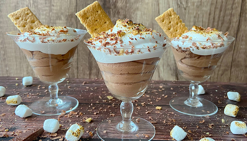 Three glass dishes of s’mores chocolate mousse sit on a wooden surface. Marshmallows and chocolate are scattered around them.