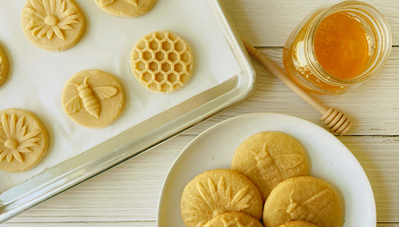 Des biscuits sont posés sur une plaque de cuisson et une assiette blanche sur une surface blanche. À côté d'eux, un pot de miel et un bâton de miel sont visibles.