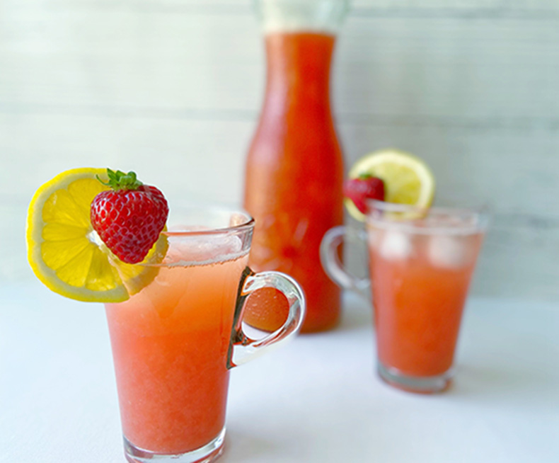 A tall glass of bright pink lemonade stands on a counter in the foreground. In the background, another glass and a carafe filled with lemonade are visible. Lemon wedges and strawberries decorate the rims of the glasses.