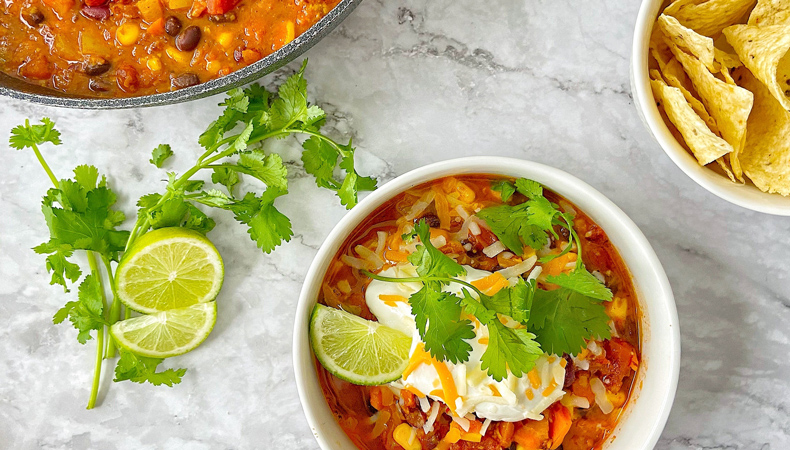 An aerial view shows a bowl of chili, garnished with sour cream, cilantro, lime, and tortilla chips, on a marble countertop. A pot of chili, a bowl of tortilla chips, and some loose garnishes are also visible.