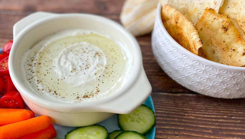 Deux petits bols blancs sont posés sur une surface en bois sombre ; l'un est rempli de sauce à la feta et l'autre de chips de pita. Des carottes, des poivrons et des concombres coupés en morceaux sont visibles autour du bol de sauce.