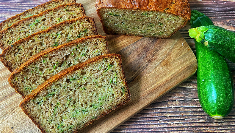 Des tranches de pain aux courgettes et un demi-pain sont posés sur un plateau en bois. Une courgette est posée à côté du pain.