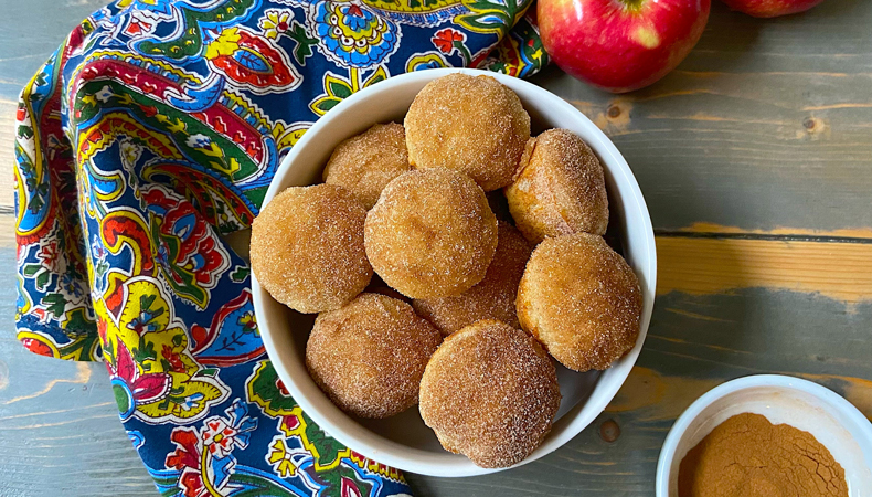 Un bol de muffins recouverts de sucre est posé sur une surface en bois. Une serviette colorée, des pommes et un plat de cannelle sont visibles.