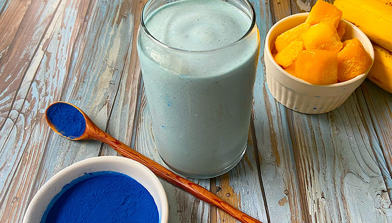 Un verre rempli d'une boisson bleue épaisse est posé sur une surface rustique en bois. Un bol et une cuillère contenant de la poudre bleue se trouvent au premier plan, avec des mangues et des bananes à l'arrière-plan.