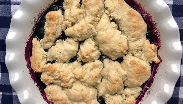 Un cobbler aux bleuets dans une assiette à tarte ronde blanche repose sur une nappe à carreaux bleue et blanche.