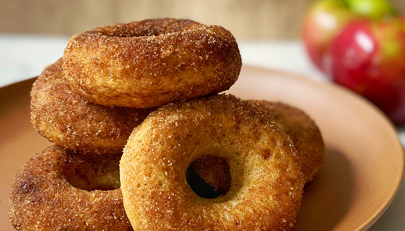 Une assiette est remplie de beignets ; des pommes sont visibles à l'arrière-plan.