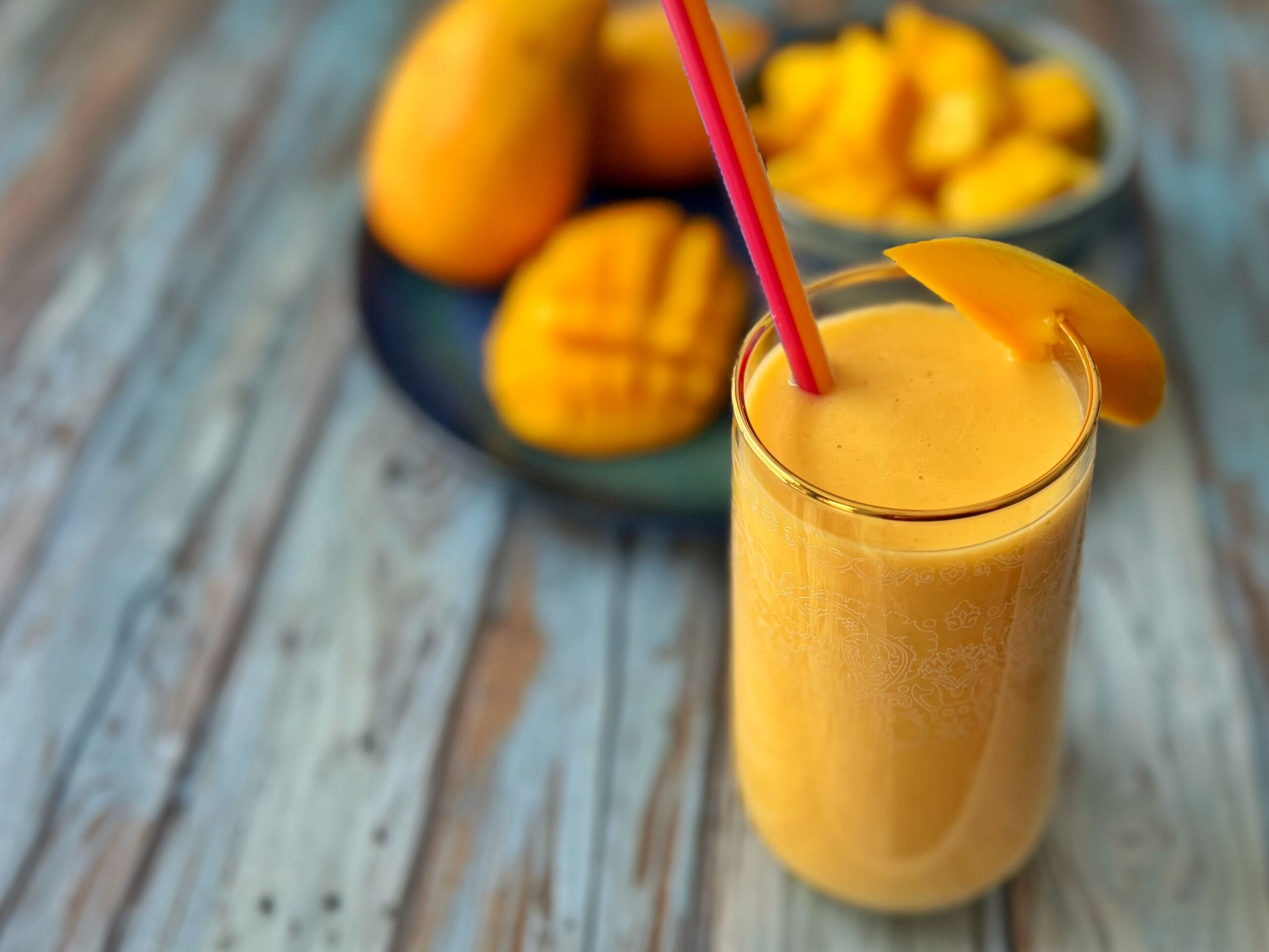 A creamy orange drink in a glass with a red straw. A plate of mangoes are visible in the background.