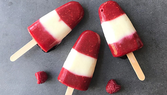 Three red-and-white striped popsicles rest on a grey surface, along with two strawberries.