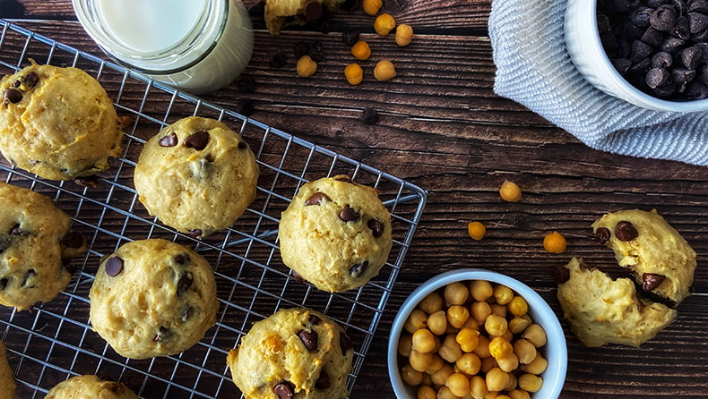 Une vue aérienne montre plusieurs biscuits sur une grille de cuisson, contre une surface en bois sombre. À côté de la grille, deux petits bols blancs sont remplis de pois chiches et de pépites de chocolat.