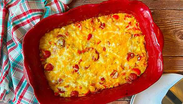 An aerial view depicts a casserole inside a red serving dish. A plaid tea towel and a metal serving utensil are visible next to the dish.