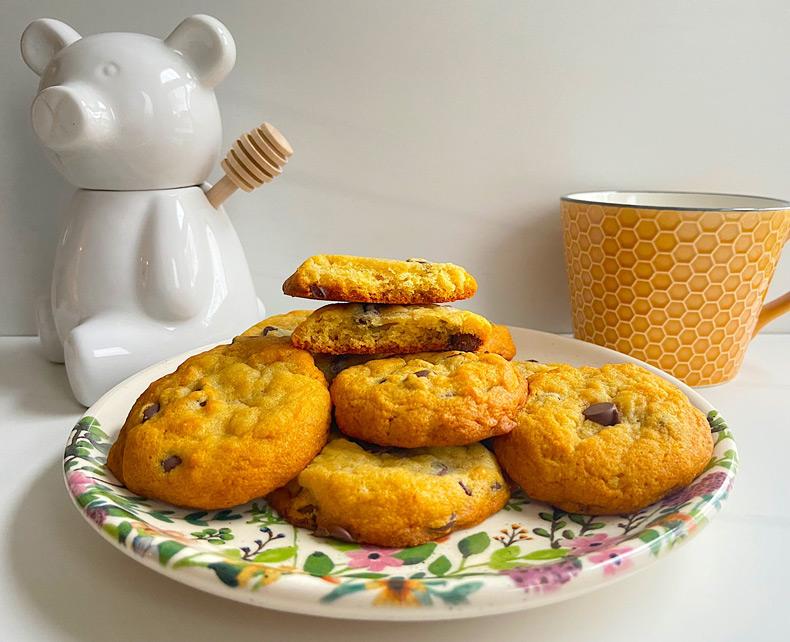 Une pile de biscuits aux pépites de chocolat est posée sur une assiette colorée. Un récipient à miel en forme d'ours et une tasse avec un motif en nid d'abeille sont visibles à l'arrière-plan.