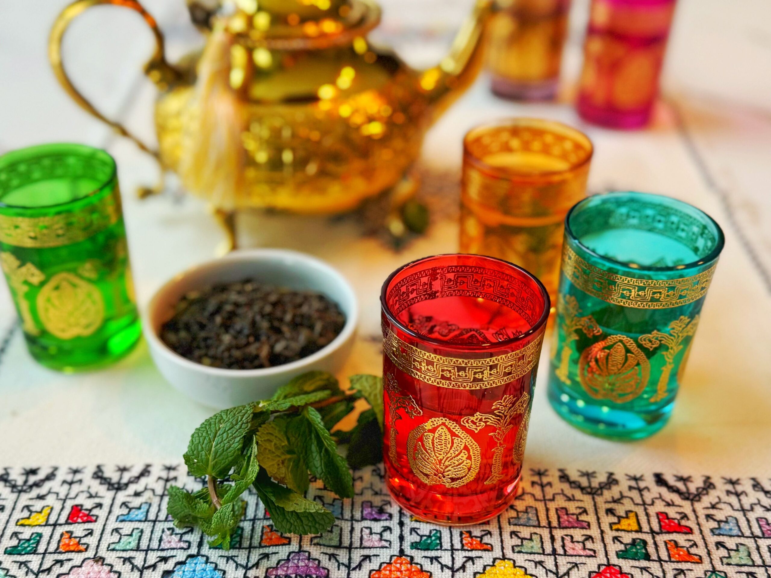 Colourful glasses of tea with green mint leaves in the forefront and a golden teapot in the rear of the photo