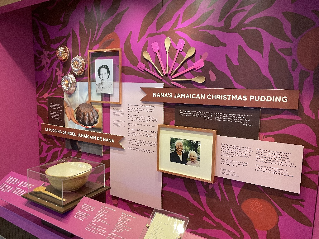 Photograph of an exhibition display, which features photographs and cooking instruments to make 'Nana's Jamaican Christmas pudding'. The display features a bright pink graphic backdrop.