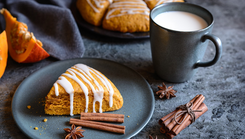 Un scone est posé sur une assiette, avec des bâtons de cannelle et des épices. En arrière-plan, on aperçoit une autre assiette de scones, des tranches de citrouille et une tasse de lait.