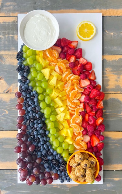 Une vue aérienne d’un plateau rectangulaire blanc, couvert d’un assortiment de fruits frais aux couleurs de l’arc-en-ciel. Un petit plat dans le haut contient une trempette de yogourt blanche et un deuxième plat dans le bas contient plusieurs bouchées d’avoine.